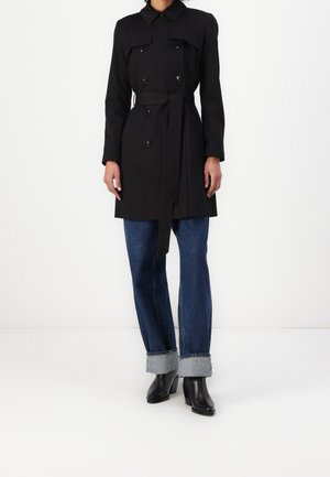 Femme portant un trench-coat noir ceinturé, un jean bleu foncé retroussé et des bottines noires, debout devant un fond blanc.