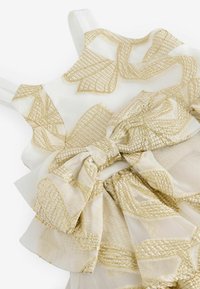 Il vestito presenta un tessuto bianco decorato con motivi geometrici dorati e un grande fiocco in vita, realizzato in materiale morbido e trasparente.