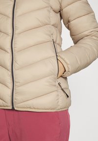 Beige pufferjakke med horisontalt kviltet mønster, lynlås lukning og sidelommer. Materialet virker glat og let skinnende.