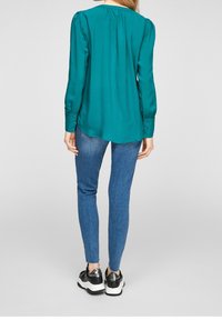 Blusa di raso teal con maniche lunghe, polsini con bottone e schiena arricciata. Abbinata a jeans blu e sneakers nere con dettagli in argento.