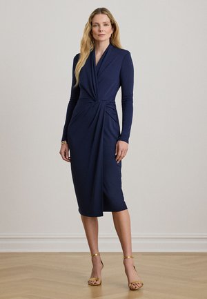 Kobieta o długich blond włosach, w granatowej sukience midi z długim rękawem i złotych sandałach na wysokim obcasie, stojąca na drewnianej podłodze.