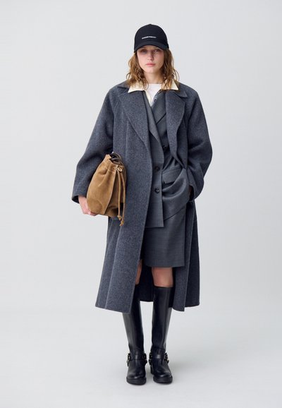 Abrigo de lana gris con solapas anchas, superpuesto a un traje gris. Botas de cuero negras hasta la rodilla y un bolso de ante marrón en forma de cubo. Gorra negra.