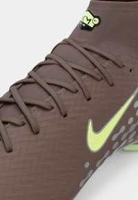 Κοντινό πλάνο καφέ ποδοσφαιρικού παπουτσιού Nike με λαχανί σήμα, μοτίβο στη σόλα και δεμένα κορδόνια πάνω σε υφασμάτινο πάνω μέρος με υφή.