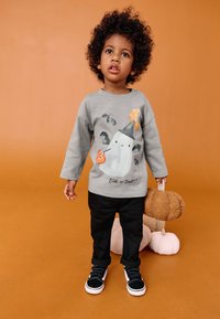 Szara koszula z długim rękawem z grafiką ducha, czarne spodnie i czarne buty. Dziecko trzyma pluszową dynię o teksturowanej powierzchni. Pomarańczowe tło.