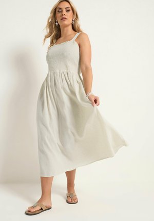 Kobieta w bezrękawowej, jasnobeżowej sukience midi z marszczonym stanem i sandałach, pozująca na jednolitym, neutralnym tle.