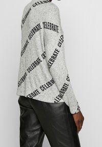 Grå tröja med svart "CELEBRATE"-text tryckt diagonalt, långa ärmar, avslappnad passform, uppvisad med svarta läderbyxor.