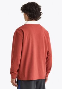 Camicia a maniche lunghe color terracotta, con colletto bianco e vestibilità rilassata. Tessuto liscio con un'etichetta vicino all'orlo.