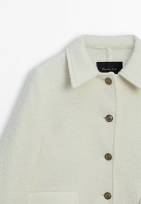 Manteau en laine blanche texturée avec un col, six boutons métalliques et poches latérales. Étiquette visible à l'intérieur du col.