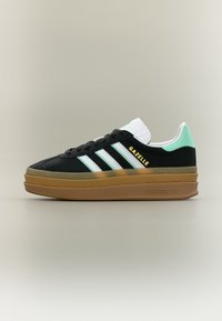 Zapatilla negra con rayas blancas y verde menta, talón blanco texturizado y suela de goma marrón. Presenta la marca "Gazelle" en dorado.