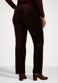 Osoba v tmavě hnědých rovně střižených kalhotách a vínové svetr s dlouhým rukávem, v kombinaci s hnědými botami na podpatku do kotníků, zobrazená zezadu.