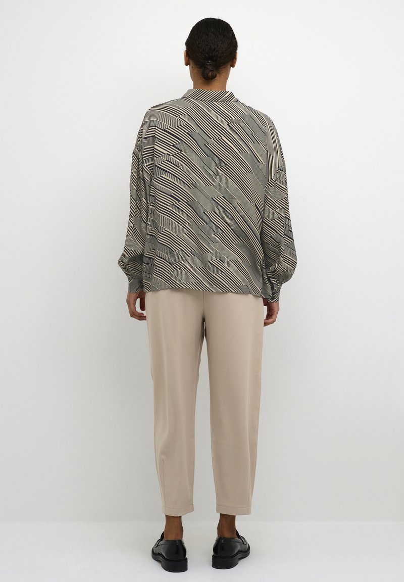 Langärmlige Bluse in einem gedämpften grünen und beigen abstrakten Muster, kombiniert mit beigen schmal geschnittenen Hosen und schwarzen Schuhen. Glattes Material, lockere Passform.