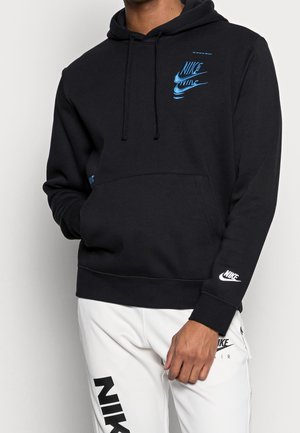 Mężczyzna w czarnej bluzie Nike z niebieskimi logo oraz białych spodniach Nike z czarnym logo, stojący na jednolitym tle.