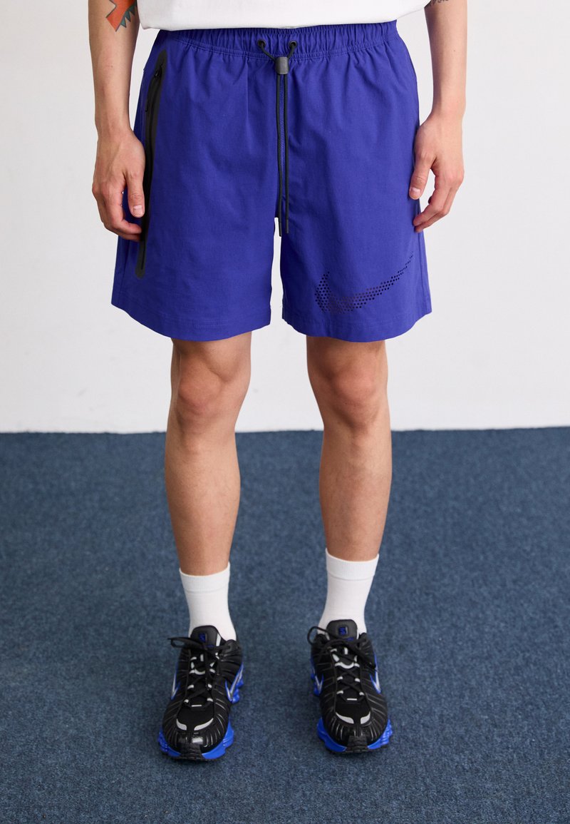 Shorts leggeri e blu con vita elastica, cordoncino regolabile, tasche con zip nere e un logo Nike in un design a punti. Indossati con scarpe nere.