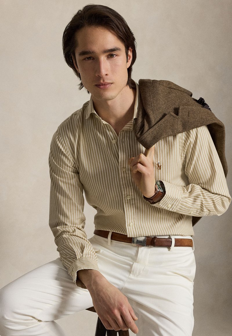 Jeune homme aux cheveux bruns, portant une chemise beige à rayures et un pantalon blanc, tenant une veste marron sur son épaule, assis sur un tabouret.