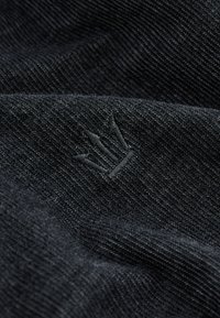Gros plan sur un tissu noir texturé avec un petit logo couronne noire à trois branches brodé au centre.