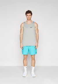 Graues ärmelloses Nike-Top, türkisfarbene Shorts, weiße Sneakers und Socken mit schwarzen Akzenten, stehend vor einem schlichten hellen Hintergrund.