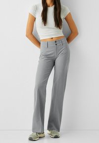 Graue weite Hosen mit glatter Textur, ausgestattet mit einem doppelten Knopfverschluss und vorderen Taschen, kombiniert mit einem taillierten grauen Oberteil und Sneakers.