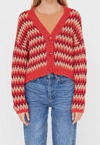 Cardigan en tricot à motif zigzag rouge et crème avec un décolleté en V, une coupe courte et trois boutons à l'avant. Jean en denim bleu classique.