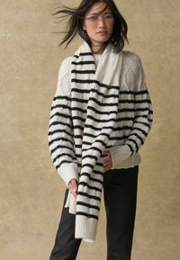 Pull en tricot épais crème avec des rayures horizontales noires, présentant un ourlet et des poignets côtelés. Accompagné de pantalons noirs, coupe décontractée.