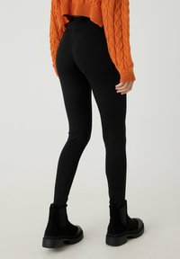 Persona con leggings ajustados negros, botas gruesas negras y un suéter de punto trenzado naranja, de pie con la espalda parcialmente girada.