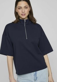 Sudadera de manga corta azul marino con cuello alto y cremallera frontal. Tejido suave, corte relajado y dobladillo recto. Pantalones cortos de mezclilla de lavado claro.
