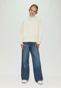 Pull en maille crème à col roulé avec des accents côtelés, accompagné d'un jean en denim foncé à jambes larges et de baskets blanches.