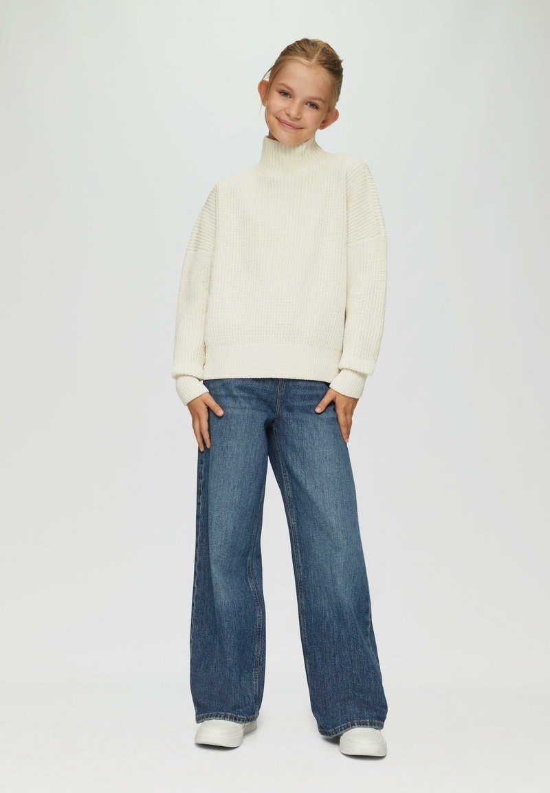 Pull en maille crème à col roulé avec des accents côtelés, accompagné d'un jean en denim foncé à jambes larges et de baskets blanches.