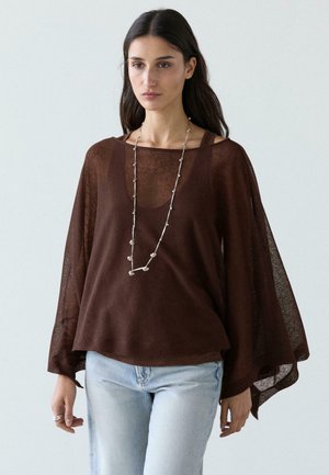 Donna che indossa una maglia marrone leggera e trasparente sopra una canotta, abbinata a jeans azzurri, una lunga collana d'argento e anelli sulla mano sinistra.