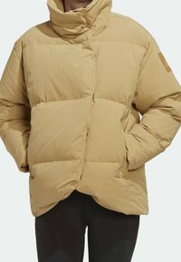 Veste matelassée beige avec col montant et fermeture éclair diagonale, portée avec les mains dans les poches et pantalon noir visible en dessous.