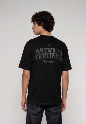 Junger Mann mit lockigem Haar steht mit dem Rücken zur Kamera und trägt ein schwarzes T-Shirt mit dem Text "MIXED FEELINGS" auf dem Rücken in dunkelgrauen Buchstaben.