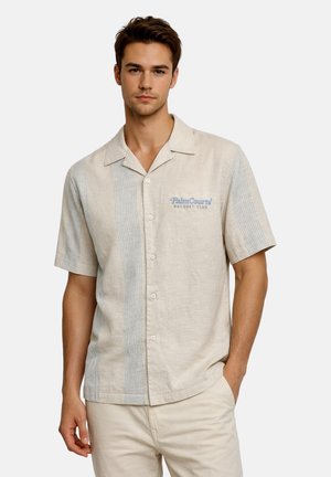 Junger Mann mit kurzem braunem Haar, der ein beiges Kurzarmhemd mit hellblauen vertikalen Streifen und dem Logo "Palm Courts Racquet Club" trägt.