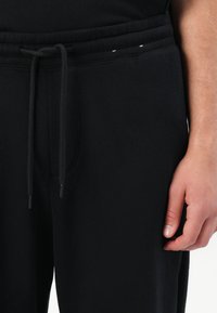 Čierne tepláky vyrobené z jemného materiálu s elastickým pásom, nastaviteľnou šnúrkou a bočnými vreckami. Majú minimálne švy.