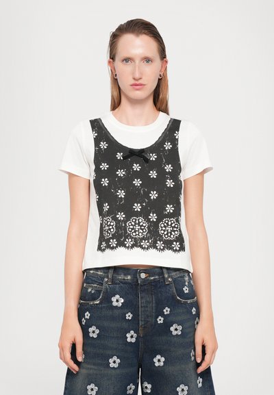 T-shirt bianco cropped con sovrapposizione di pizzo nero caratterizzato da ritagli floreali e dettaglio a fiocco, abbinato a pantaloni corti in denim scuri decorati con fiori ricamati.