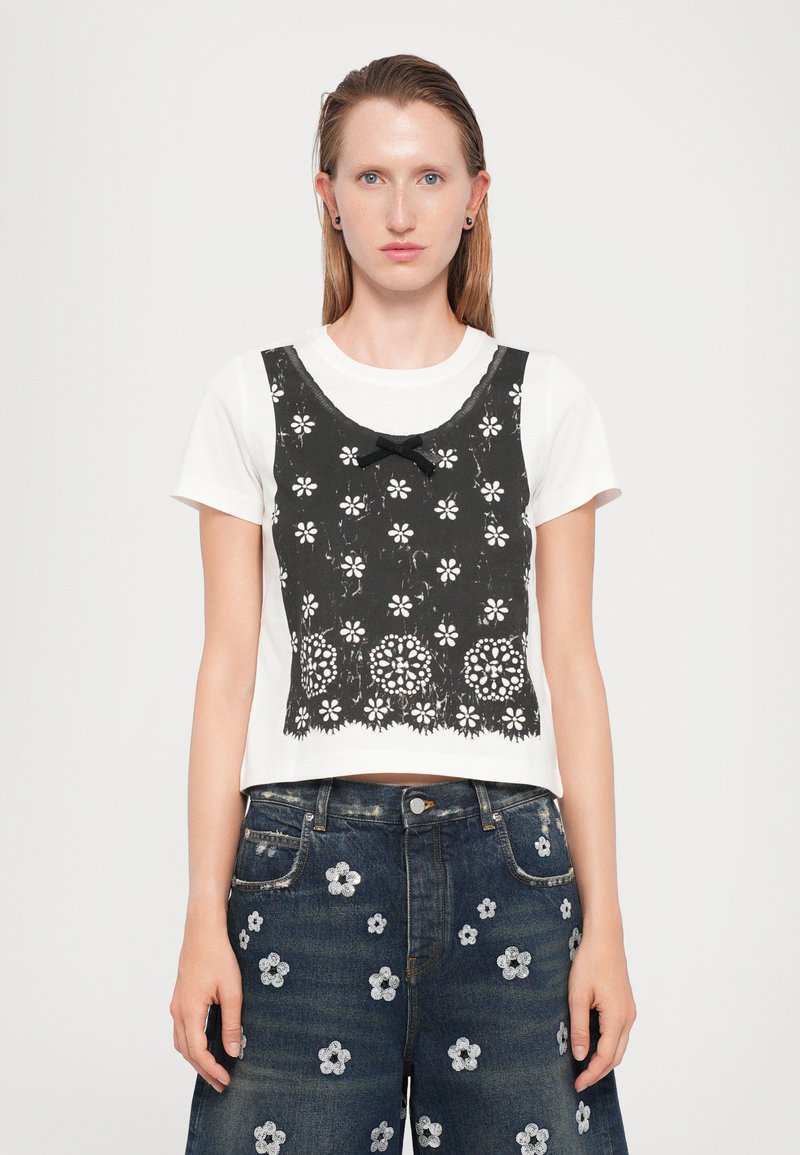 Geknipte witte t-shirt met zwarte kant overlay, voorzien van bloemvormige uitsparingen en een strikdetail, in combinatie met donkere denim shorts versierd met geborduurde bloemen.