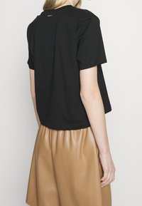 Camiseta negra corta con mangas cortas, con cuello redondo y un pequeño detalle de etiqueta en la parte de atrás, combinada con una falda brillante beige.