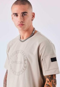 Camiseta de algodón beige con logo embellecido circular, mangas cortas y un parche negro texturizado en la manga izquierda.