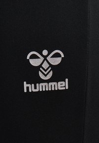 Logo d’abeille stylisé blanc avec le texte « hummel » en dessous sur un fond de tissu noir texturé.