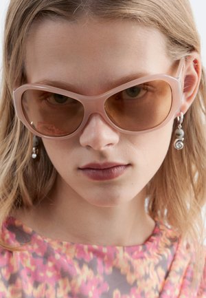 Jeune femme aux cheveux clairs porte de grandes lunettes de soleil teintées de rose, des boucles d'oreilles spirales argentées et un top rose floral.