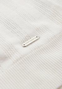 Tejido de punto blanco con un patrón texturizado y una etiqueta plateada que lleva el texto "QUINTESSENCE", mostrando puños acanalados y una apariencia general suave.