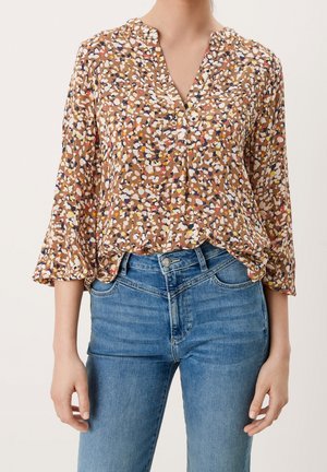 Bruine blouse met meerkleurig abstract patroon en driekwart mouwen, gecombineerd met hooggetailleerde blauwe spijkerbroeken bij een staande persoon.