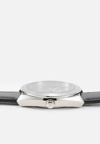 Reloj plateado con una esfera redonda, correa de cuero negra, bisel delgado y una corona en el lateral para ajustes. Cubierta de vidrio transparente.
