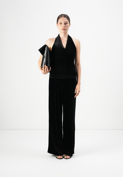 Vrouw in een zwart haltertop en wijde broek met een grote zwarte clutch, staande tegen een eenvoudige witte achtergrond.