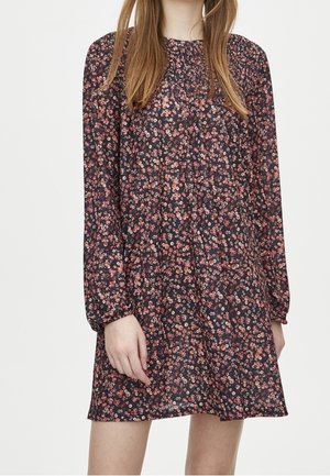 Kvinde iført en langærmet sort kjole med et lille pink og hvidt blomsterprint, stående mod en ensfarvet baggrund.