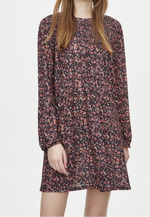 Femme portant une robe noire à manches longues avec un petit motif floral rose et blanc, debout devant un fond uni.