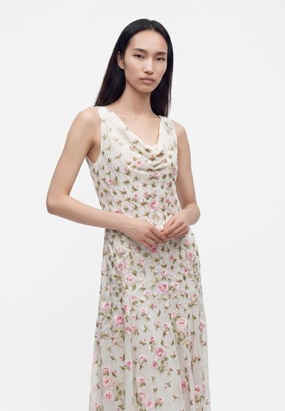 Femme aux longs cheveux noirs portant une robe blanche sans manches avec une broderie florale rose sur un fond clair uni.