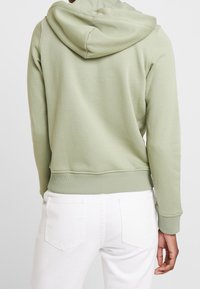 Sudadera con capucha de color verde claro, con una textura suave, costuras visibles y puños acanalados, combinada con jeans blancos que tienen bolsillos traseros.