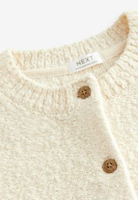Cardigan color crema realizzato in filato testurizzato, con colletto a costine, bottoni in legno marrone e un'etichetta tessuta all'interno.