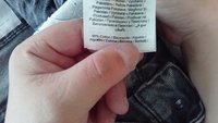 Mâini ținând o etichetă de îmbrăcăminte pe care scrie „Fabricat în Pakistan” și „99% bumbac” pe un fundal de material denim.