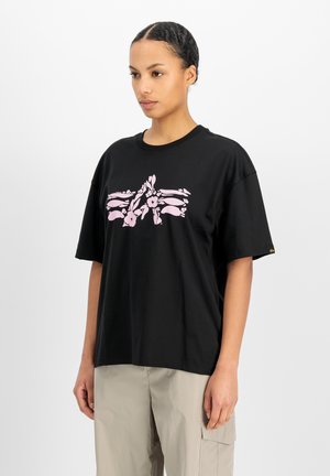 Frau trägt ein schwarzes, übergroßes T-Shirt mit pinkem Blumenmotiv und beigen Cargohosen, steht vor weißem Hintergrund.