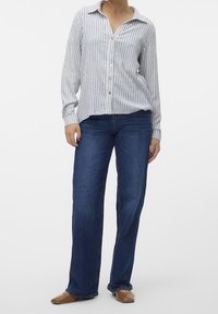 Gestreept overhemd met knopen in lichtgrijs en wit, gecombineerd met blauwe spijkerbroek. Het overhemd heeft een kraag en lange mouwen, de jeans is recht uitgesneden.